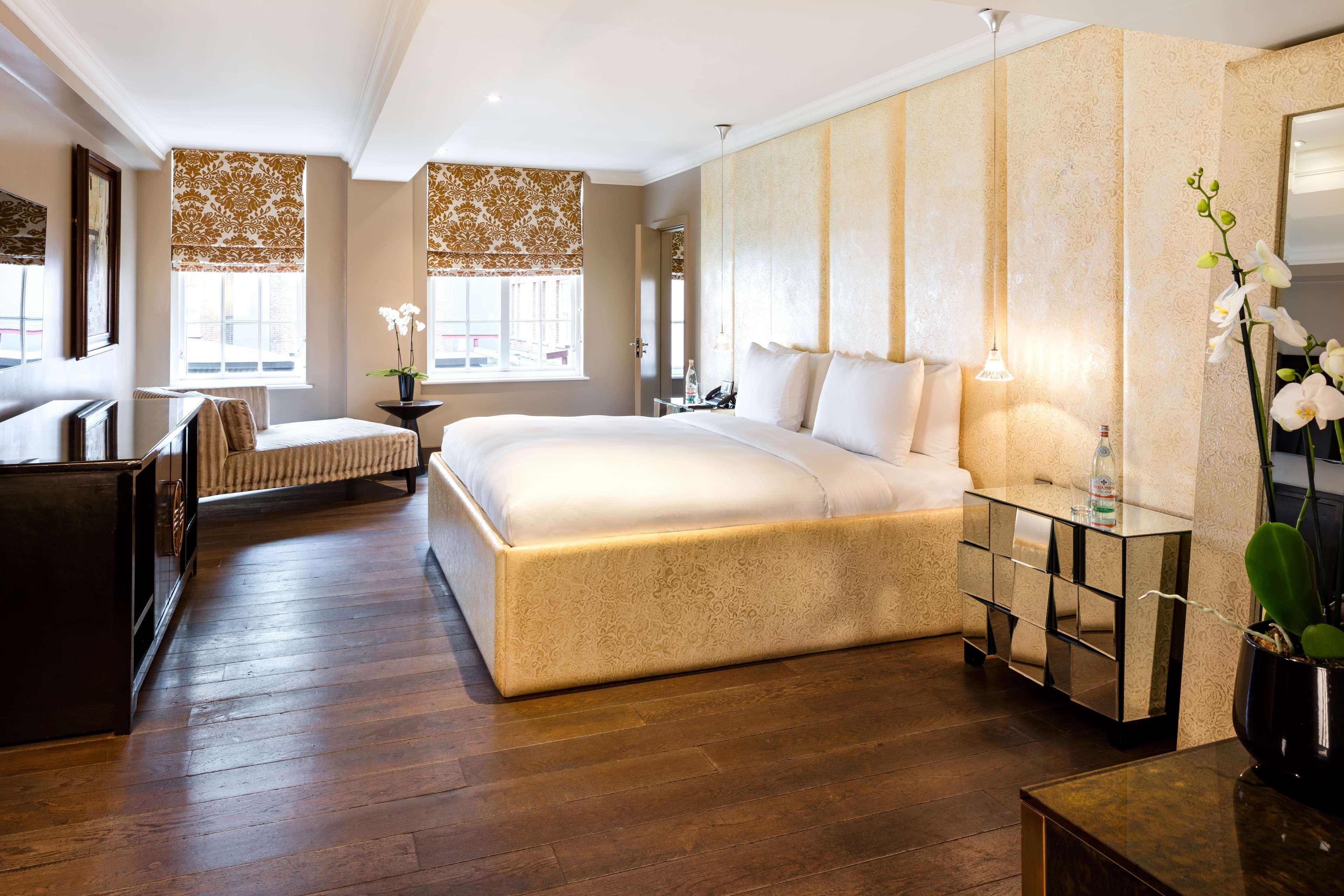 The May Fair, A Radisson Collection Hotel, Mayfair London Exterior photo The photo shows a modern and elegantly designed hotel room or bedroom. It features a large, plush bed with two pillows, situated against a light-colored wall. To one side, there is a stylish bench or chaise lounge, adding a comfortable seating area. 