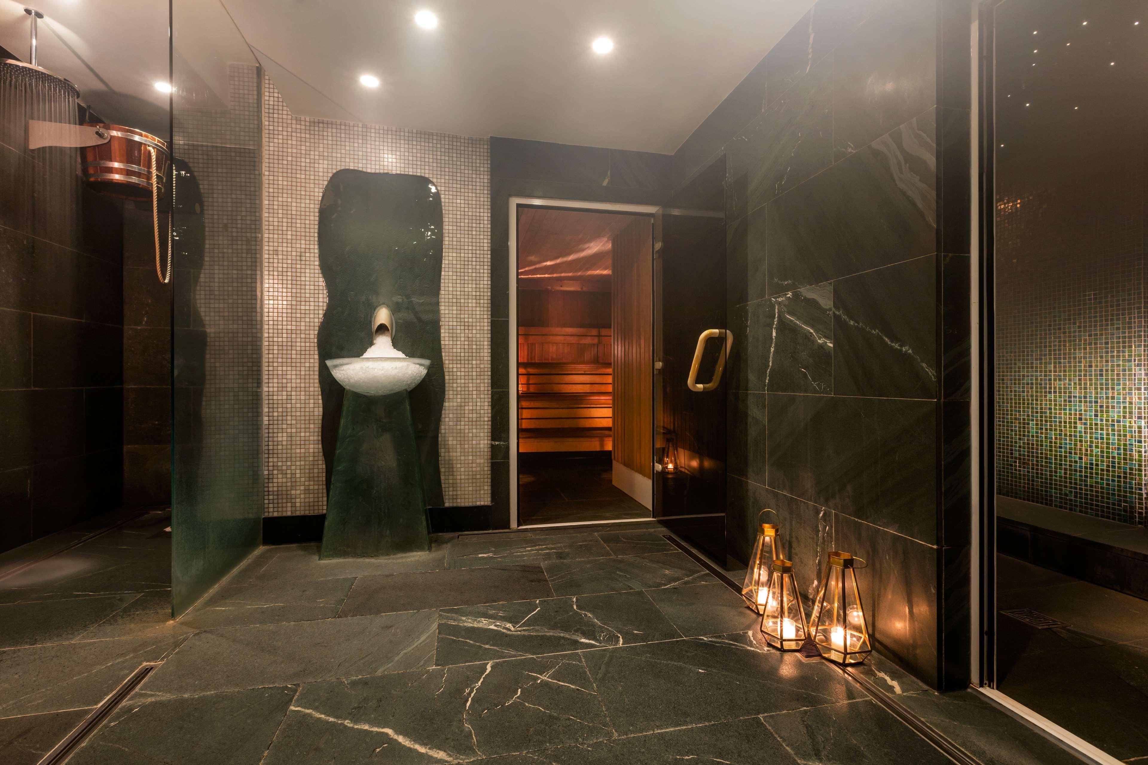 The May Fair, A Radisson Collection Hotel, Mayfair London Exterior photo The photo shows a modern and stylish bathroom or spa area. It features dark marble flooring and walls with a sleek design. On one side, there is a unique sink that has an artistic shape, mounted on the wall. Next to the sink, there is a glass door th