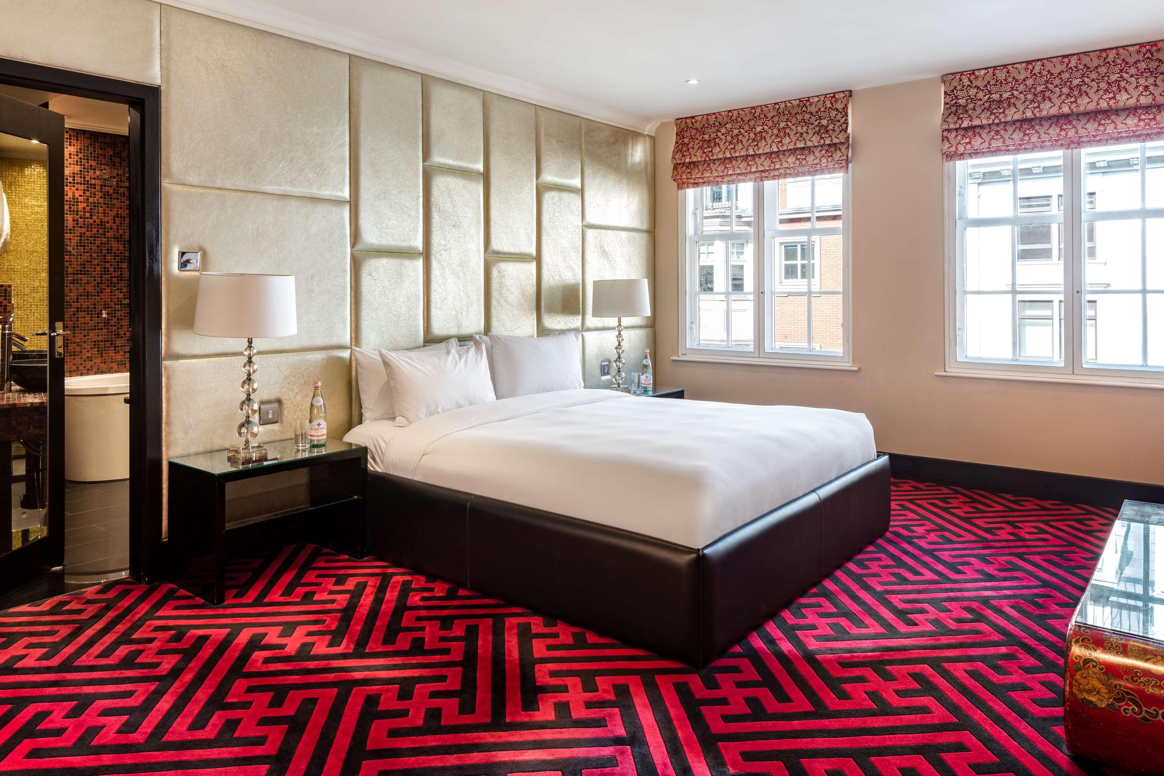 The May Fair, A Radisson Collection Hotel, Mayfair London Exterior photo The photo shows a modern hotel room featuring a large bed with white bedding. The walls are accented with a stylish, textured design, and the room is illuminated by natural light coming from two windows, which have patterned blinds. The floor is cove