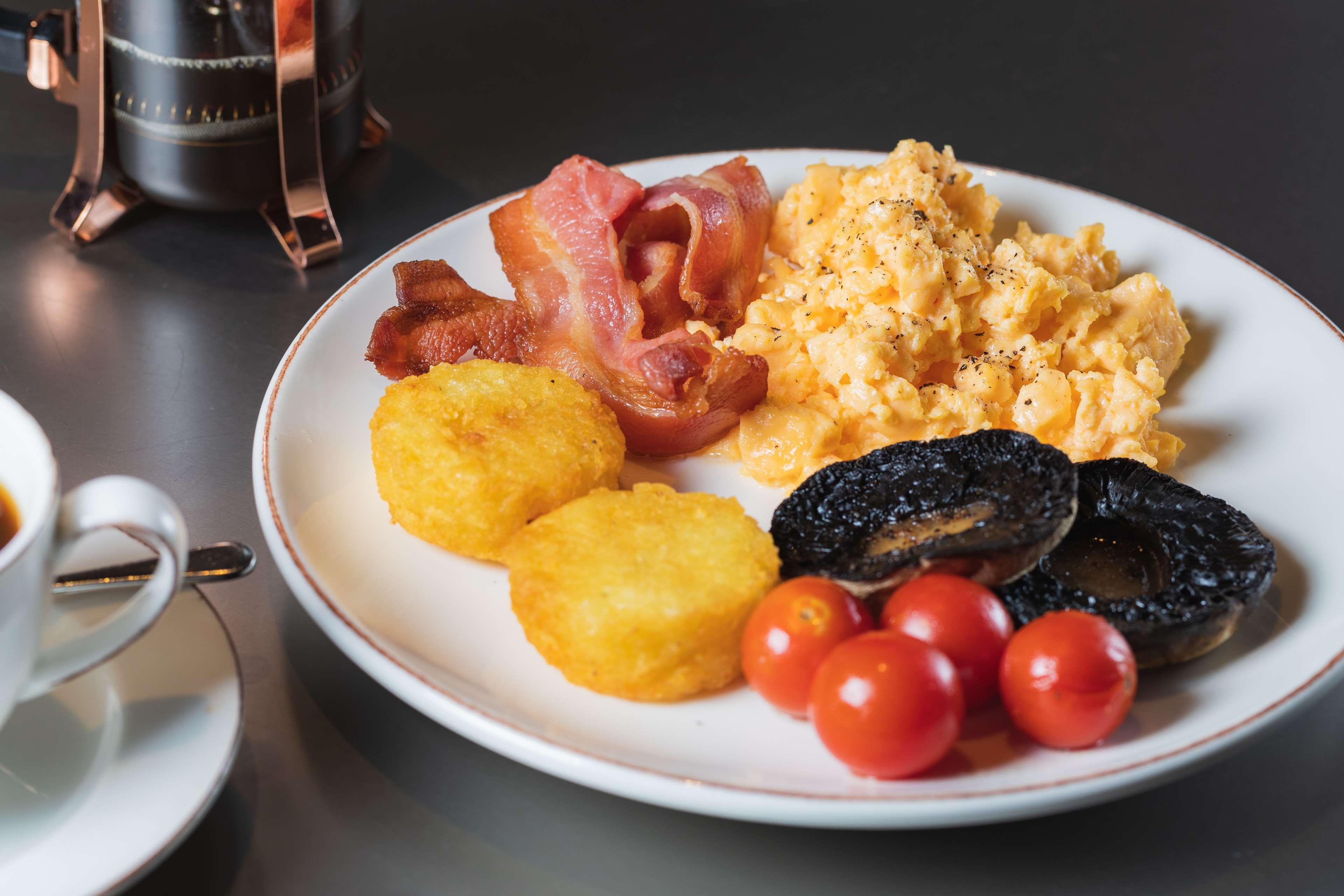 The May Fair, A Radisson Collection Hotel, Mayfair London Exterior photo The photo shows a plate of breakfast food that includes scrambled eggs, crispy bacon strips, two golden hash browns, grilled tomatoes, and slices of black pudding. The dish appears to be served alongside a small cup, presumably for coffee or tea. The