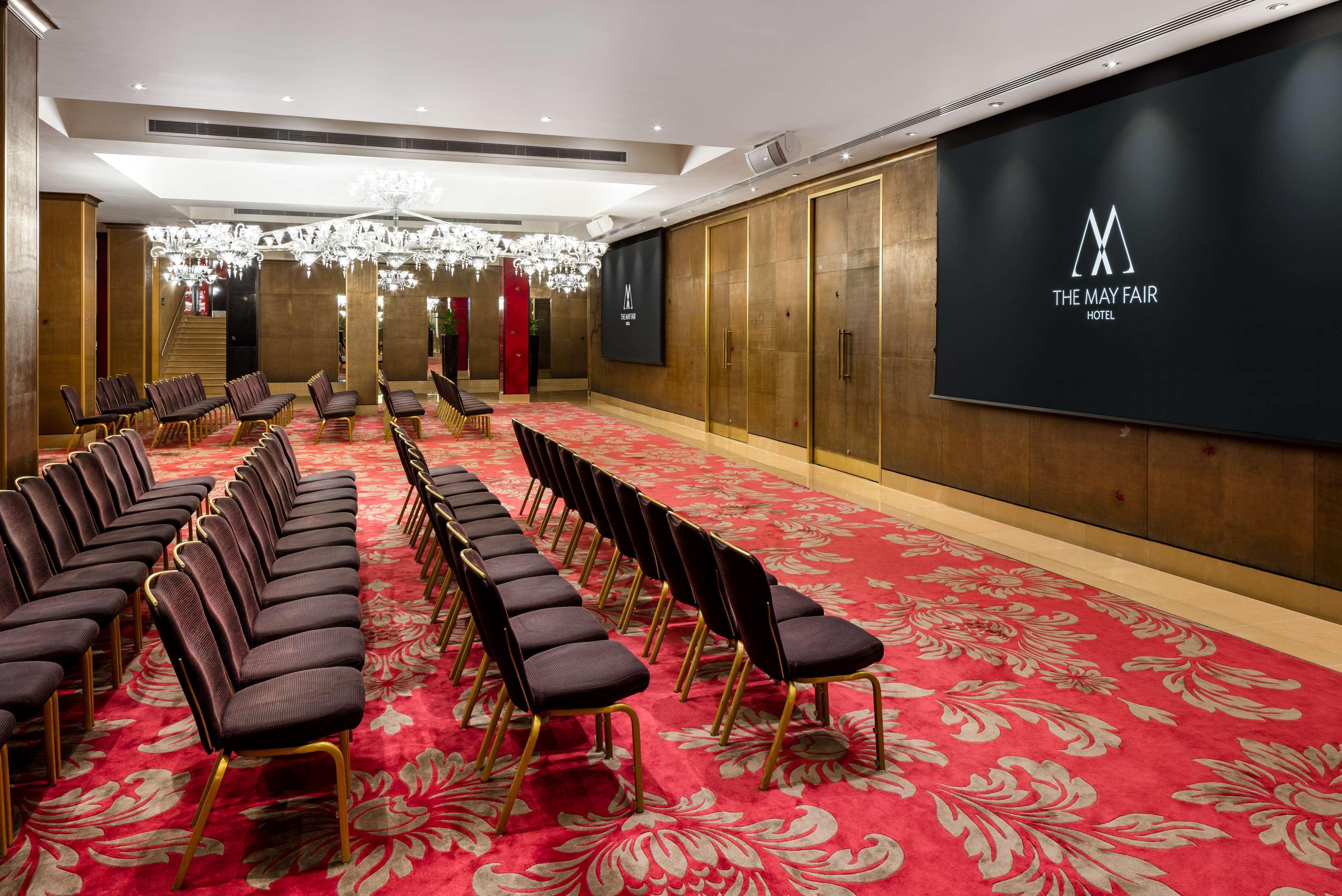 The May Fair, A Radisson Collection Hotel, Mayfair London Exterior photo The photo shows a conference or event room inside The May Fair Hotel. The space has a stylish design with decorative chandeliers hanging from the ceiling. There are rows of upholstered seating arranged in front of a large screen that displays the hot