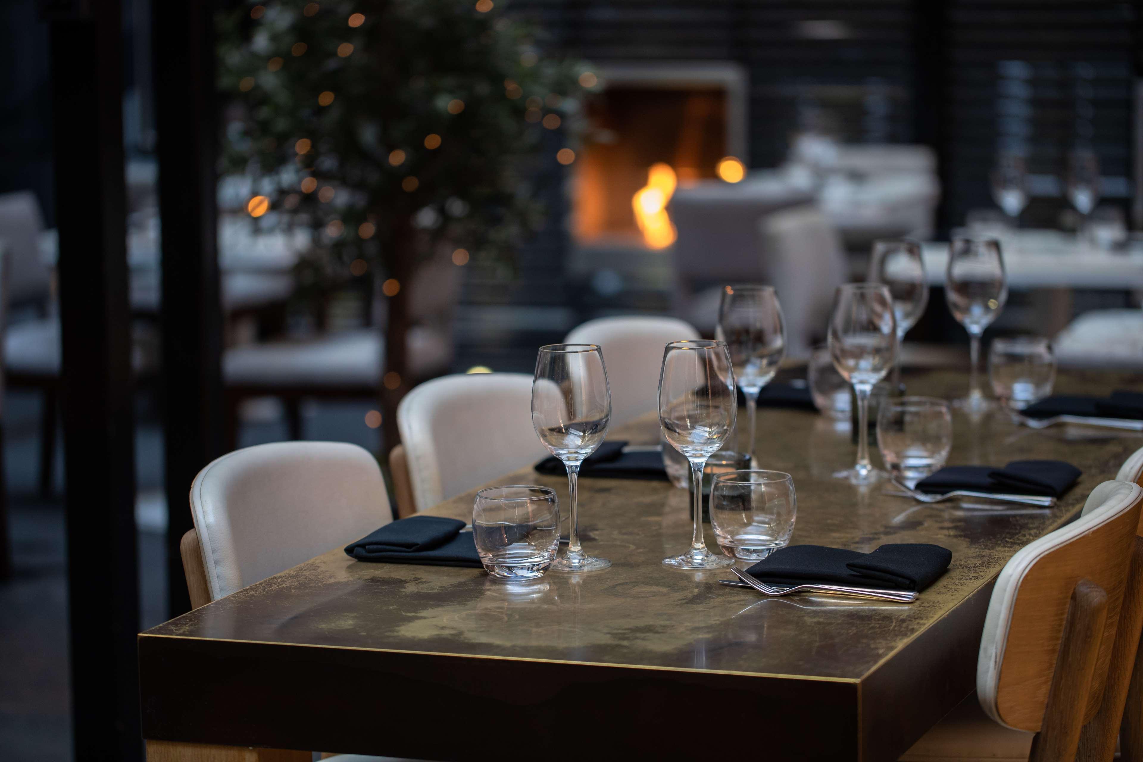 The May Fair, A Radisson Collection Hotel, Mayfair London Exterior photo The photo shows a stylish restaurant dining area. A beautifully set table is in the foreground, featuring elegant glassware and black napkins. The table has a glossy surface and appears to be well-prepared for dining. In the background, there is a co