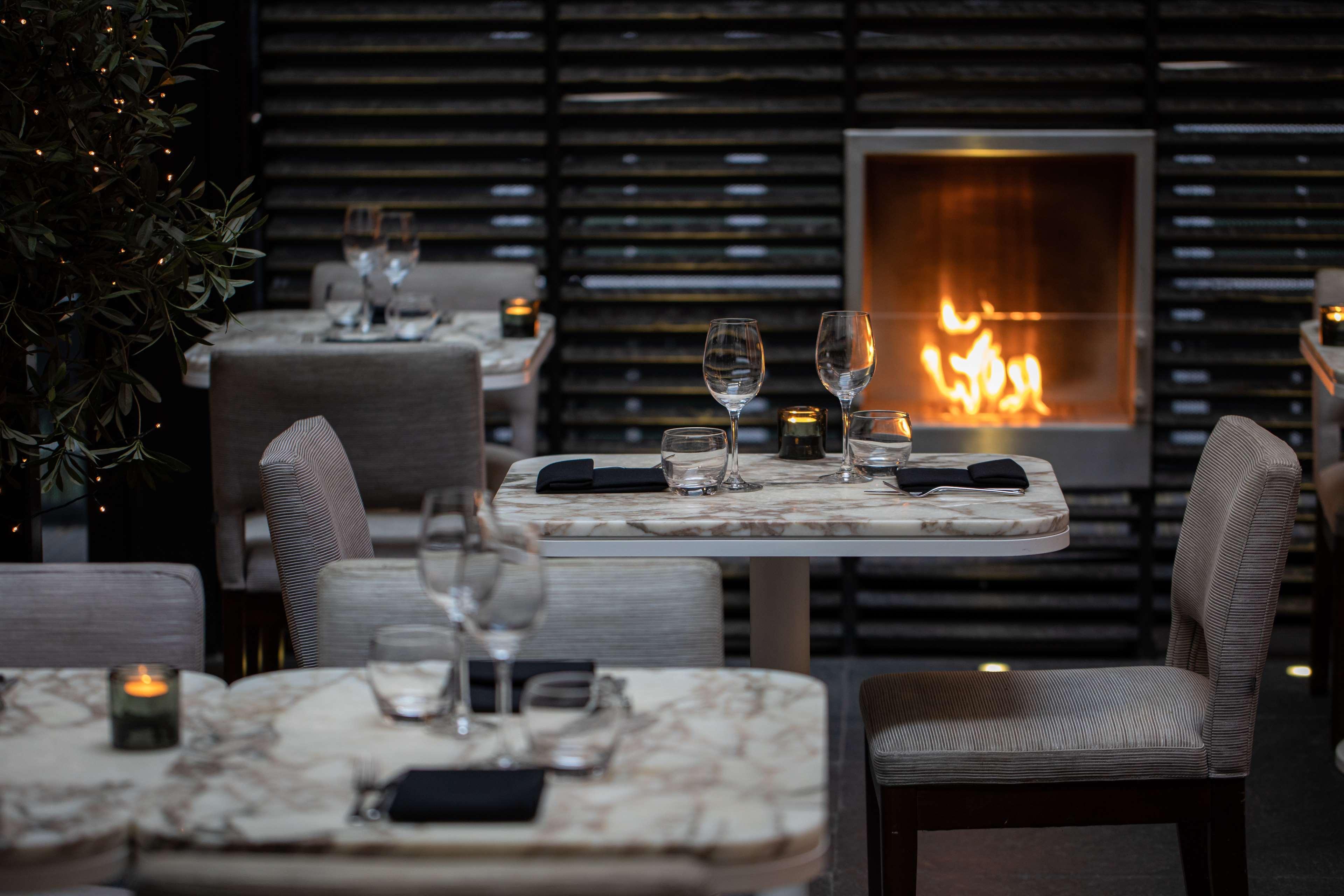 The May Fair, A Radisson Collection Hotel, Mayfair London Exterior photo The photo depicts a cozy restaurant interior with elegantly set tables. Each table features a marble top and is equipped with glassware and napkins. There are several tables neatly arranged in the space. In the background, there is a modern fireplace