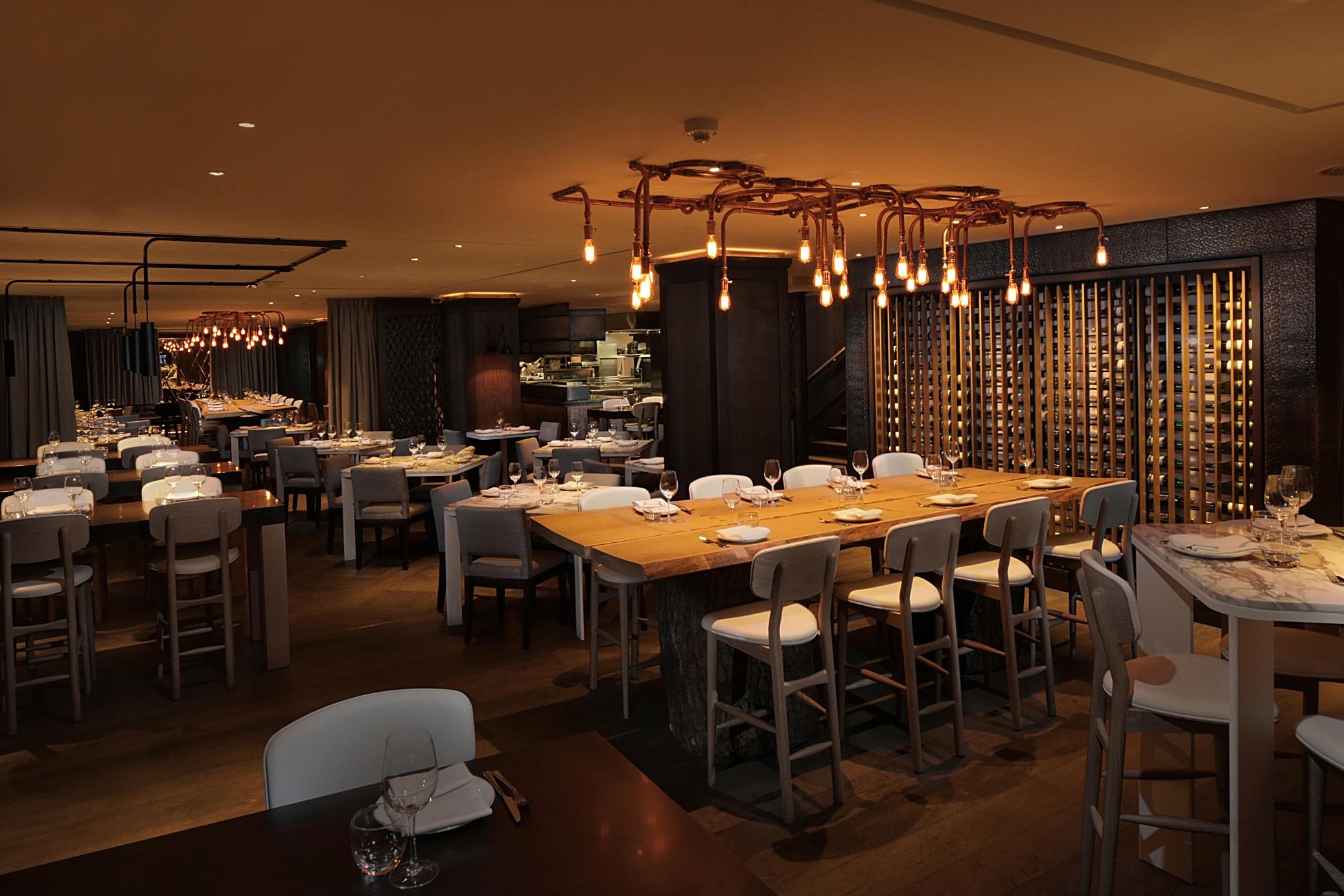 The May Fair, A Radisson Collection Hotel, Mayfair London Exterior photo The photo depicts a modern restaurant interior. The space features a combination of wooden and white furniture, with tables set for dining. There is a long, communal-style table at the center, surrounded by high chairs. The lighting is warm, coming f