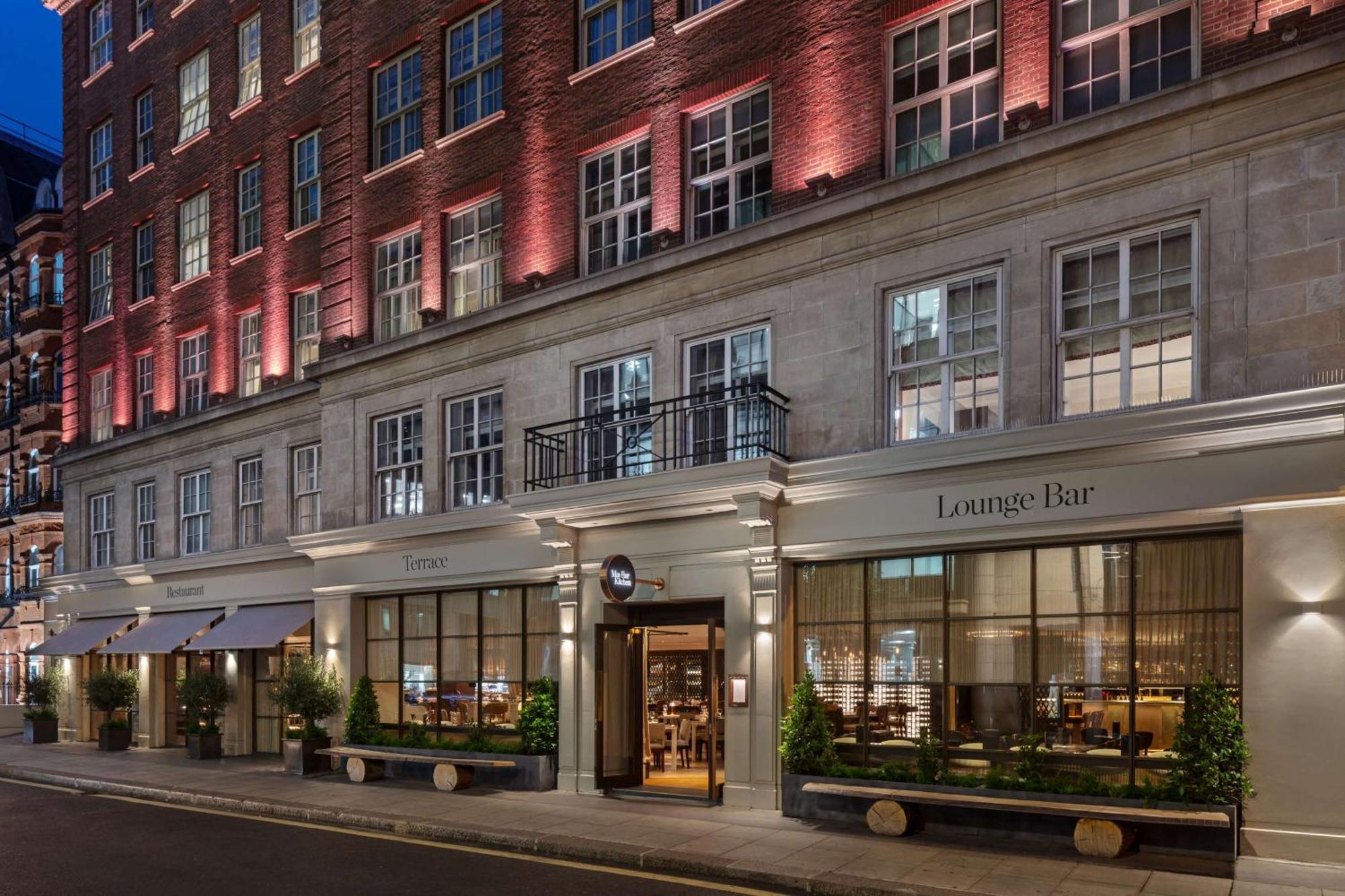 The May Fair, A Radisson Collection Hotel, Mayfair London Exterior photo The photo showcases a stylish building, likely a hotel or a high-end establishment. The facade features a mix of brick and stonework, and there are large windows that allow visibility into the lounge area. The entrance is marked by a sign indicating 