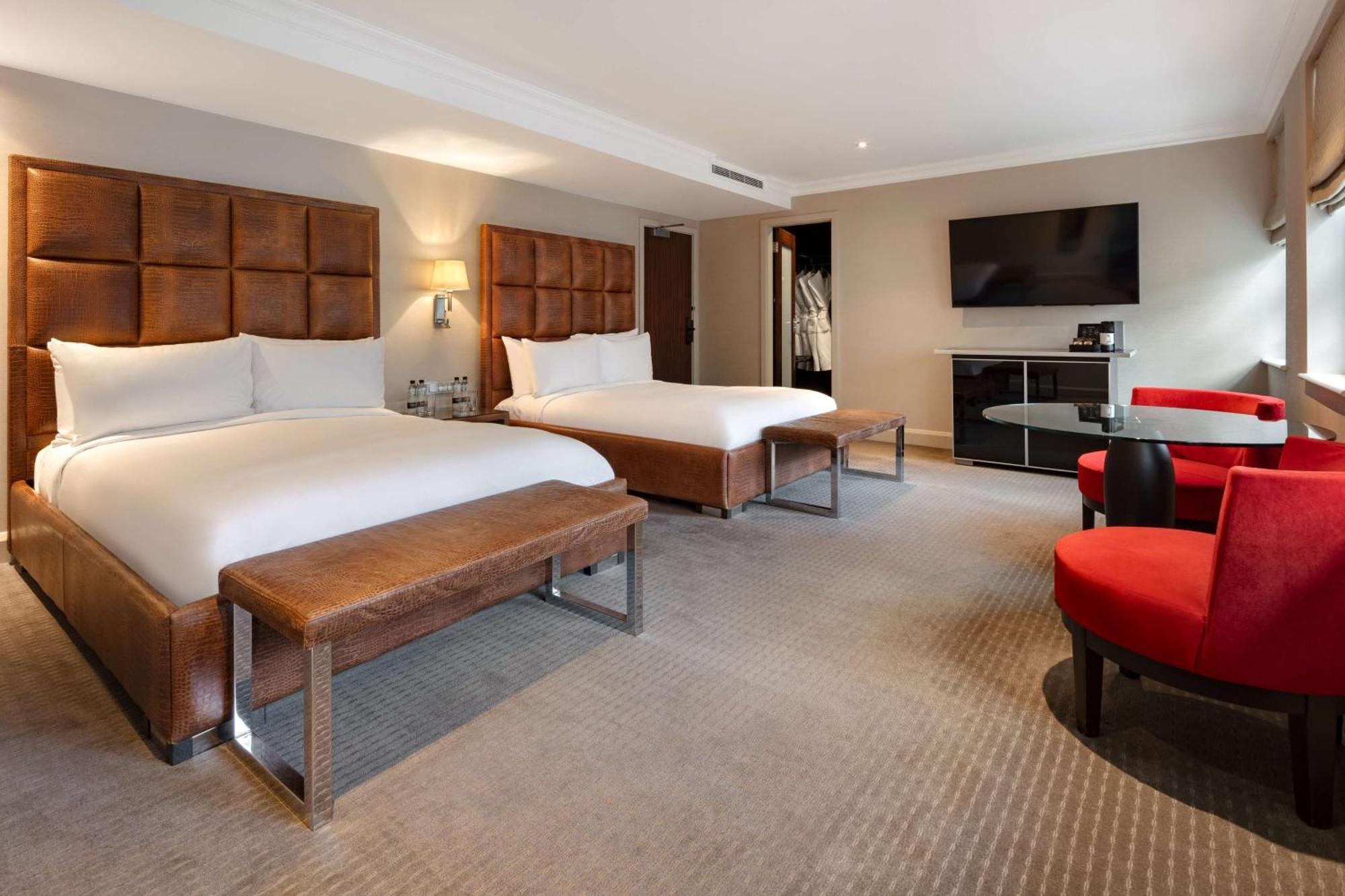 The May Fair, A Radisson Collection Hotel, Mayfair London Exterior photo The photo shows a modern hotel room with two beds. Each bed is made with white linens and has a large, upholstered headboard. There is a bench at the foot of each bed. To the right, there are two red chairs around a small glass table. A flat-screen T