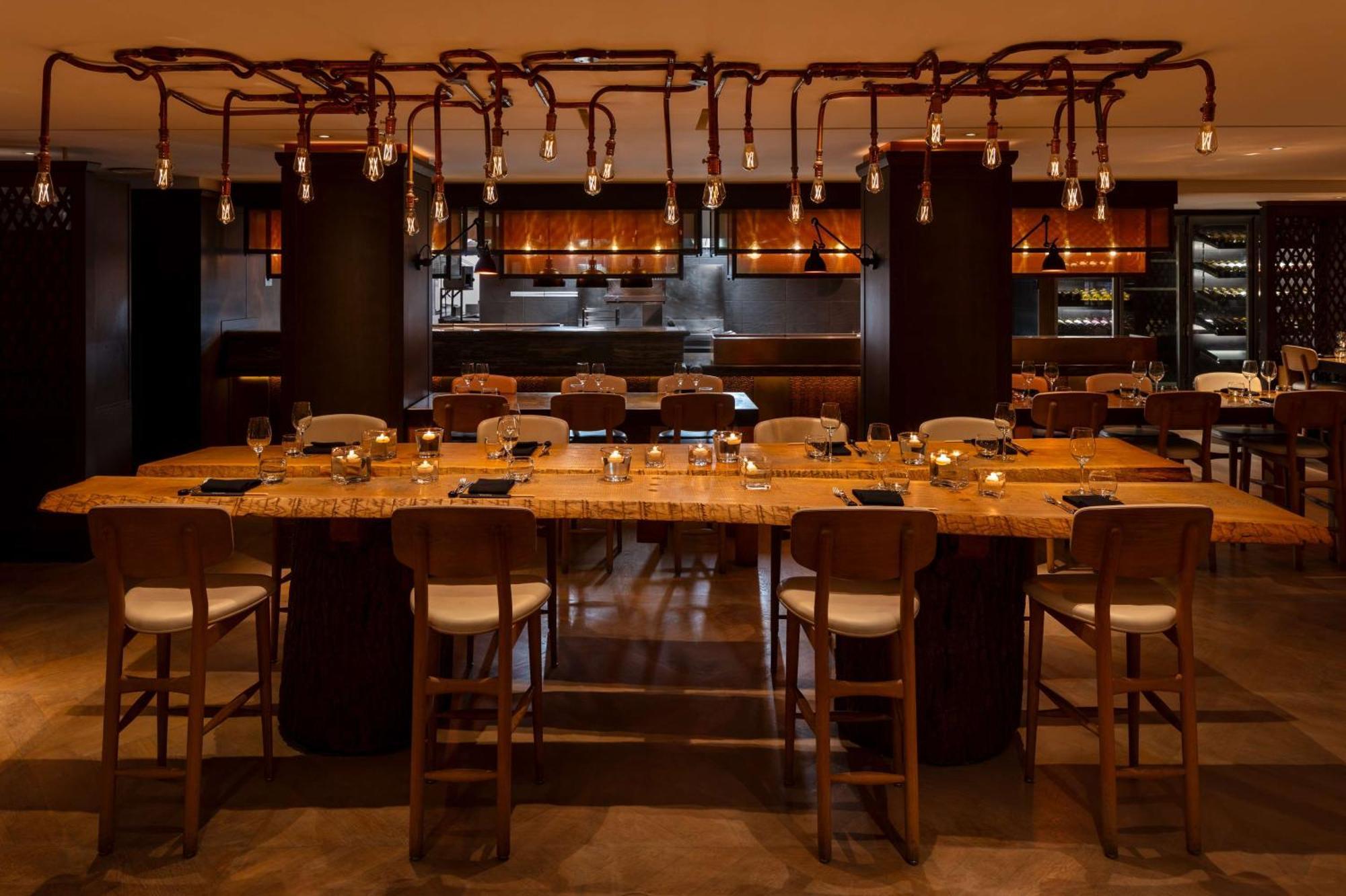 The May Fair, A Radisson Collection Hotel, Mayfair London Exterior photo The photo shows a modern restaurant interior featuring a long wooden dining table with several high chairs around it. The table is set with plates and glasses, suggesting it is ready for guests. Above the table, there are unique light fixtures that r