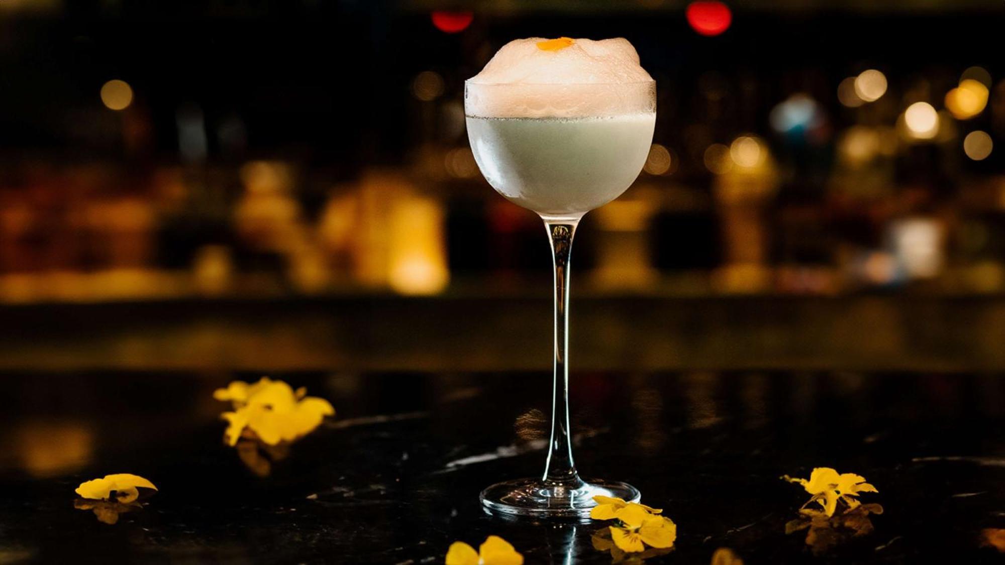 The May Fair, A Radisson Collection Hotel, Mayfair London Exterior photo The photo shows a stylish cocktail served in an elegant stemmed glass. The drink appears to be light in color, topped with a frothy layer, possibly a foam or whipped ingredient. Around the glass, there are yellow flower petals scattered on the surfac