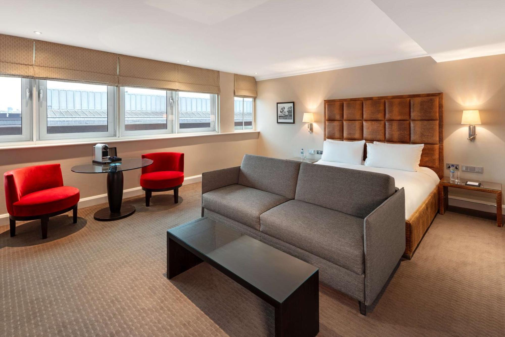 The May Fair, A Radisson Collection Hotel, Mayfair London Exterior photo The photo shows a modern hotel room featuring a comfortable setup. There is a large double bed with a headboard, some decorative artwork on the wall, and two windows allowing natural light to enter. In the room, there's a gray sofa facing a dark coff